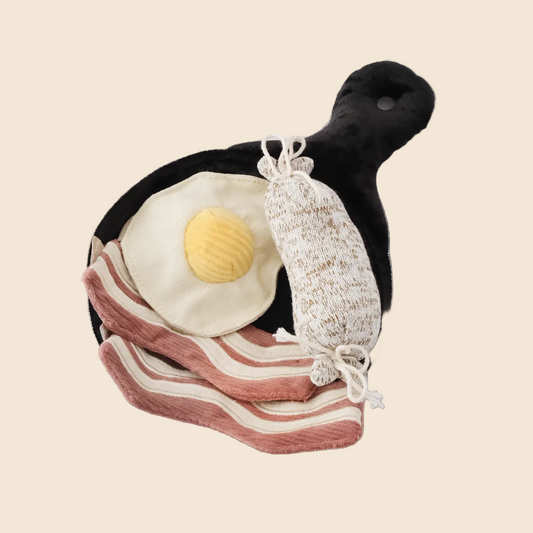 Toy set resembling a breakfast scene with a frying pan, bacon, egg, and sausage on a cream background.