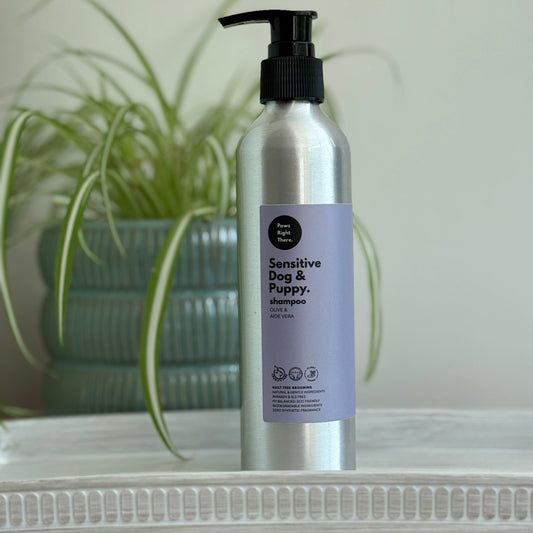 A silver pump bottle of 'Sensitive Dog & Puppy' shampoo by Happy Paws Home, standing on a white textured surface with a blurred green potted plant in the background.