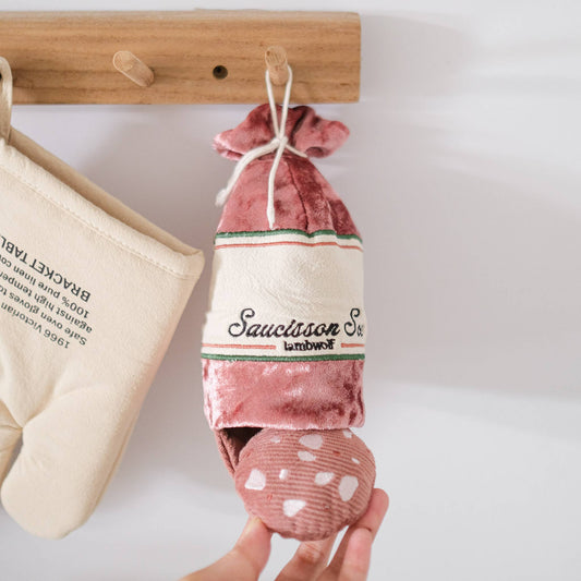 Pink plush toy shaped like a sausage with 'Saucisson Sec' text, hanging from a wooden hook.