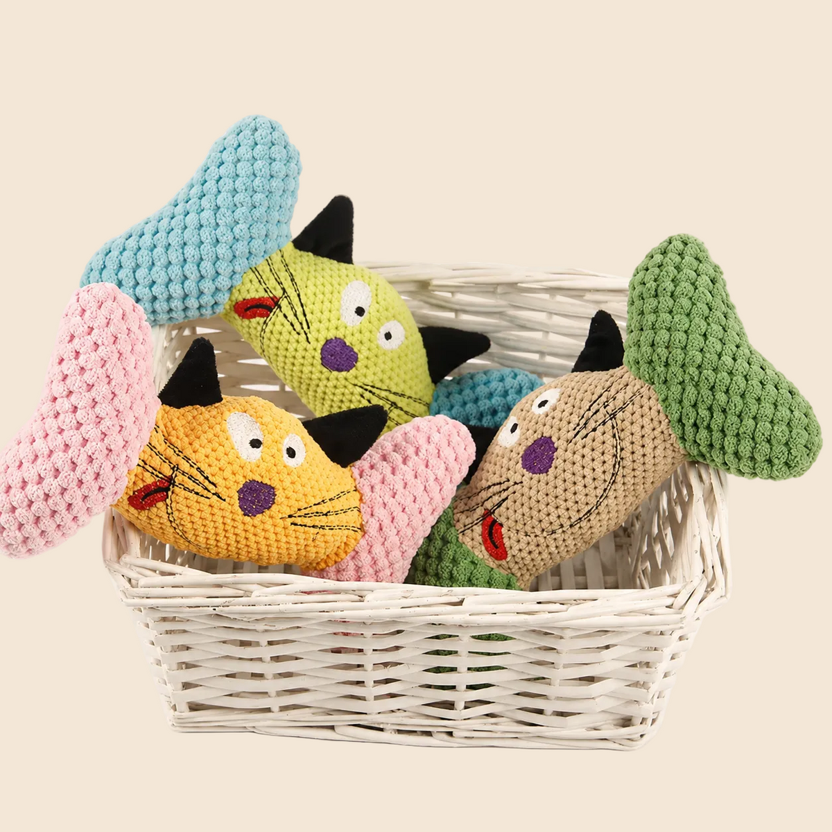 Four knitted cat-shaped dog toys in pastel colors, arranged inside a white wicker basket on a clean white background.