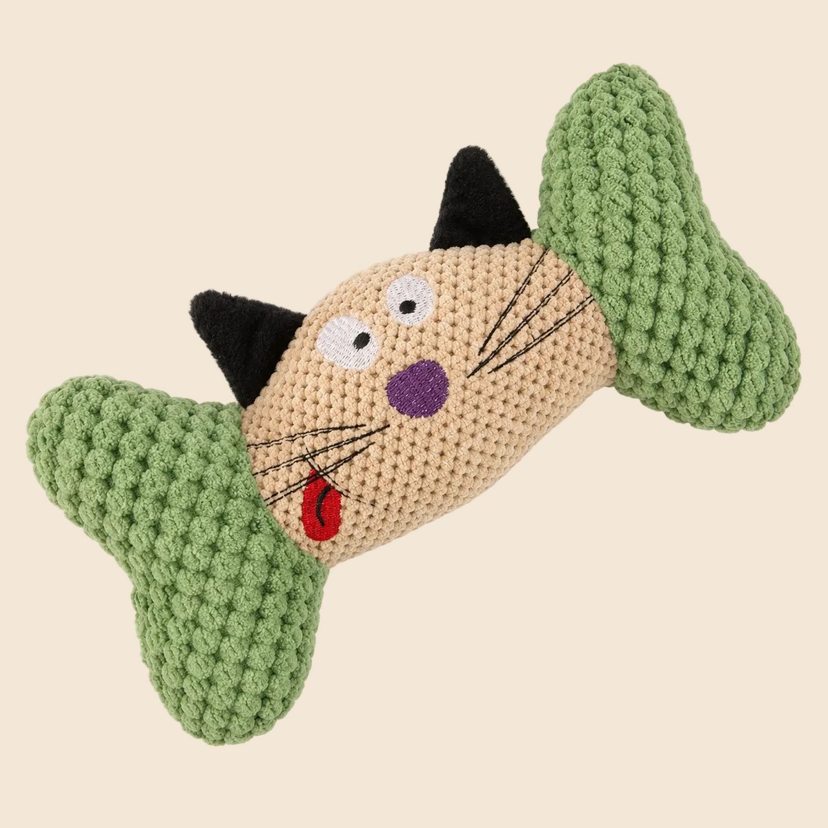 A crocheted cat-shaped pet toy with green bone-shaped ends, a beige body, black ears, and embroidered facial features including white eyes, a purple nose, and a red tongue, shown on a plain off-white background.