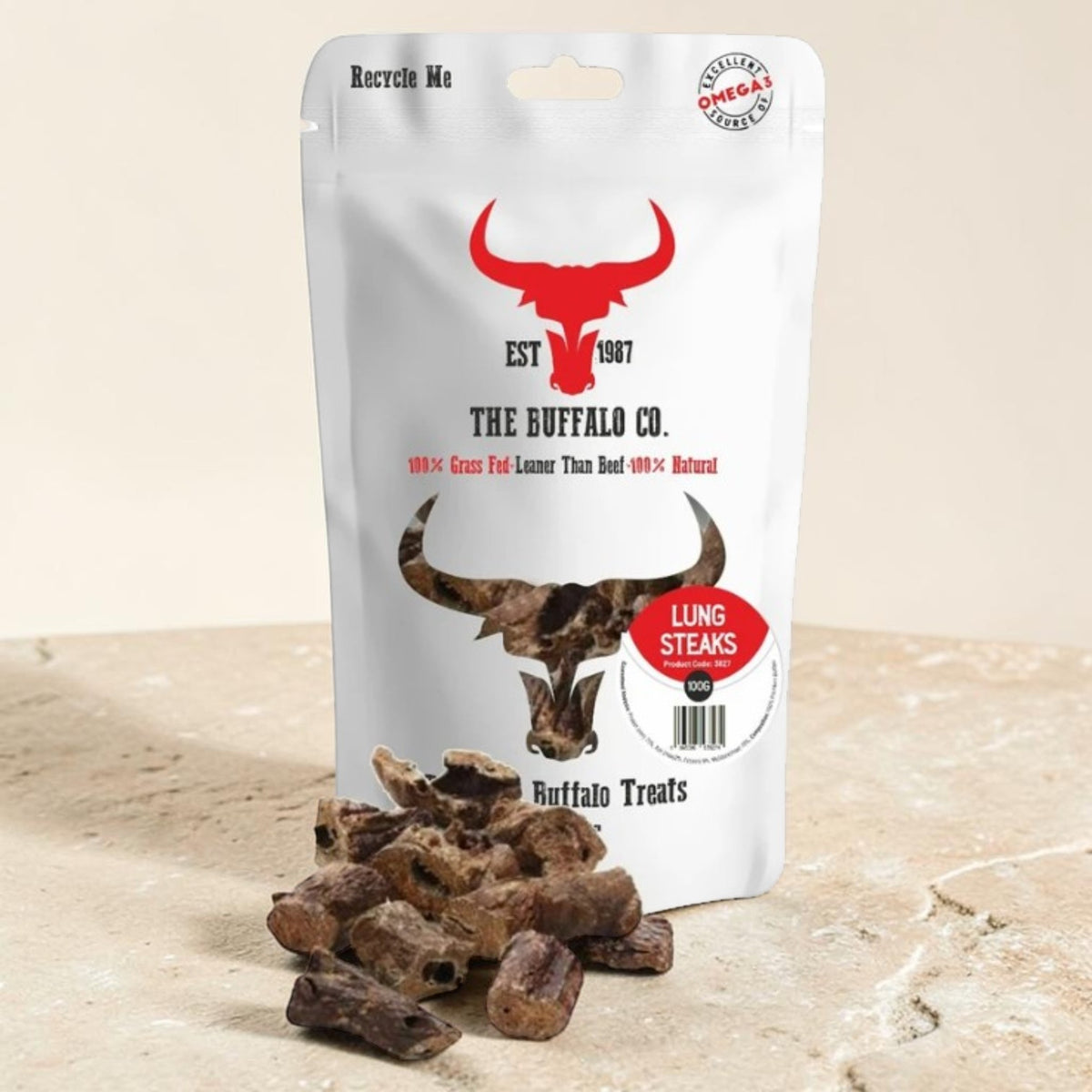 A stand-up pouch of The Buffalo Co. Lung Steaks dog treats sits on a light stone surface with several brown treat pieces placed in front.