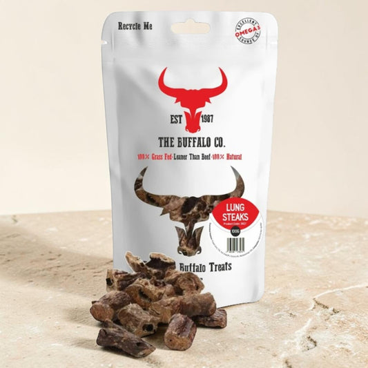 A stand-up pouch of The Buffalo Co. Lung Steaks dog treats sits on a light stone surface with several brown treat pieces placed in front.