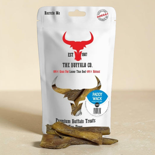 A stand-up pouch of The Buffalo Co. Paddy Wack dog treats sits on a light stone surface with several brown treat pieces placed in front.
