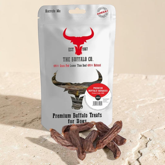 A stand-up pouch of The Buffalo Co. Premium Buffalo Treats for Dogs with a red buffalo head logo and a few brown jerky-style treats placed in front on a light stone surface.