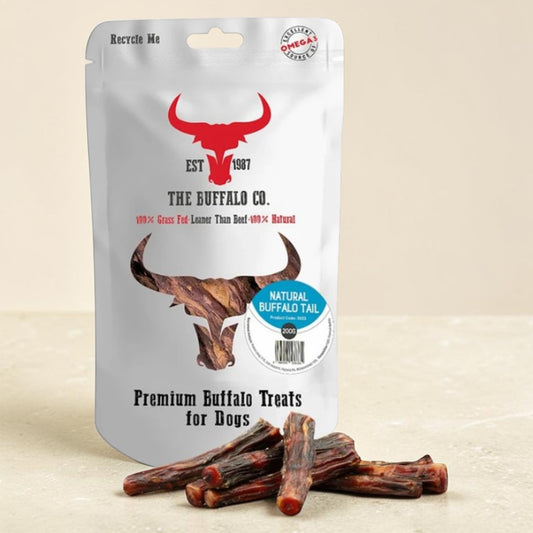 A stand-up pouch of The Buffalo Co. Premium Buffalo Treats for Dogs, with several brown buffalo tail treats displayed in front on a light beige surface.