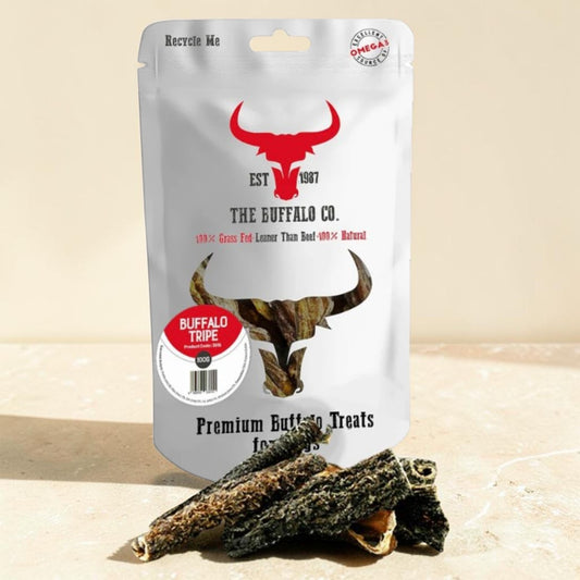 A stand-up pouch of The Buffalo Co. Premium Buffalo Treats with several dark brown jerky-style sticks placed in front on a light beige surface.