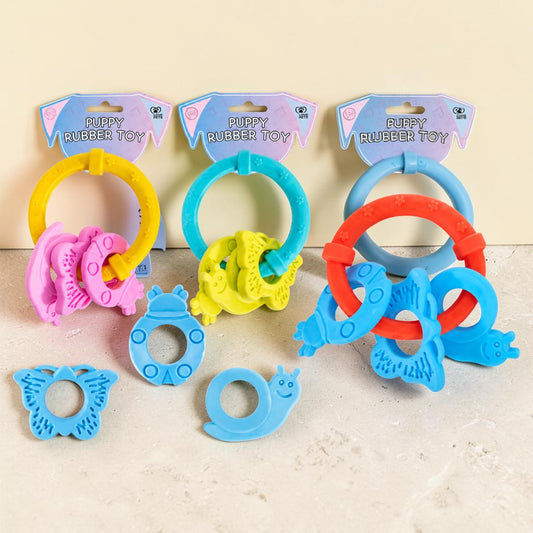 Three colorful puppy rubber teething toys in their retail packaging, displayed on a light beige surface. The toys include butterfly, snail, and elephant designs in yellow, blue, pink, and red.