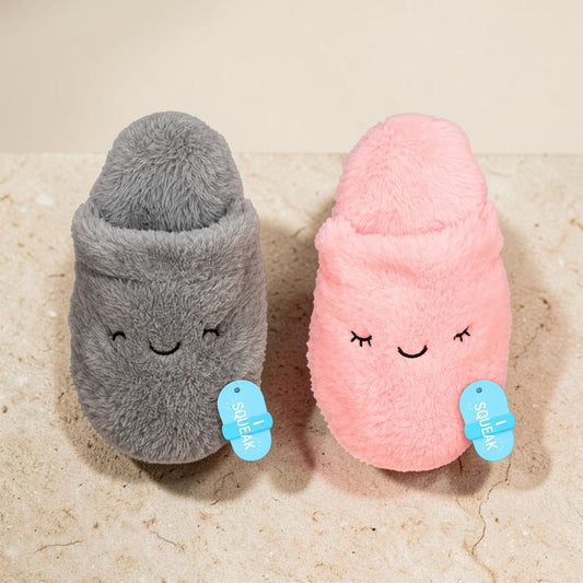 Two plush slipper-shaped toys in gray and pink, each with embroidered closed eyes and a small blue tag that reads 'SQUEAK', photographed on a light stone surface against a pale beige background.