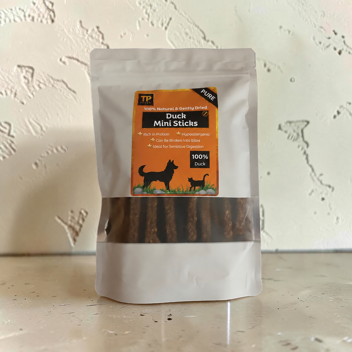 A stand-up pouch of TP Pure Duck Mini Sticks dog treats, featuring an orange label with black and white graphics, set against a textured off-white wall.