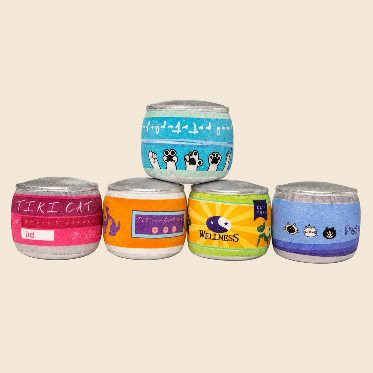 Four small cylindrical plush toy cans with silver lids arranged in a pyramid on a light cream background. The top can is light blue with black cat silhouettes and white script, the middle-left is pink with 'TIRI CAT', the middle-right is orange with a blue label, and the bottom-right is blue with black cat faces.
