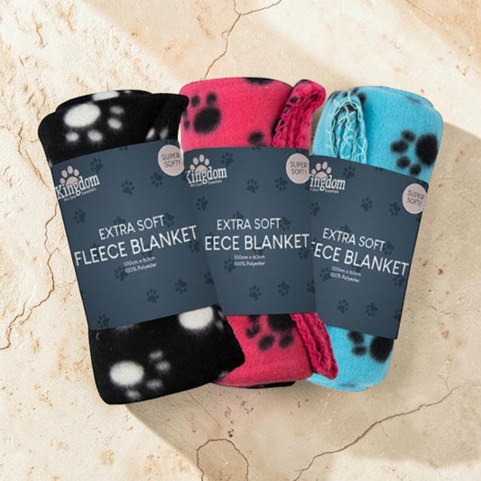 Three folded pet fleece blankets with dark blue paper bands are arranged on a light beige stone surface. The left blanket is black with white paw prints, the middle is pink with black paw prints, and the right is light blue with black paw prints.