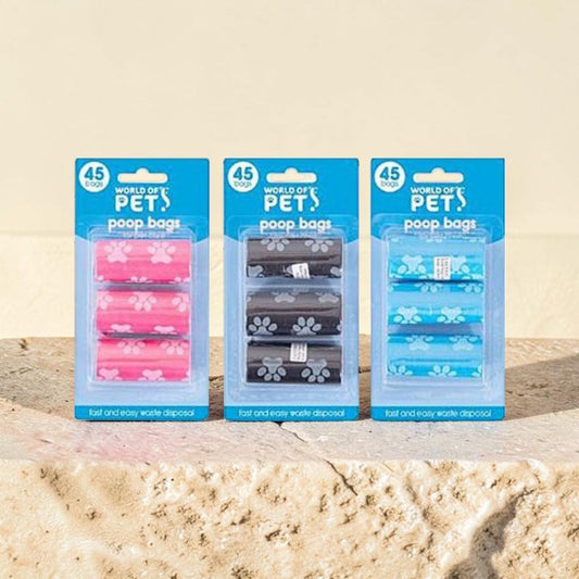 Three packages of World of Pet poop bags in pink, black, and blue, displayed on a stone surface against a beige background.