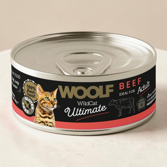 A can of Wolf WildCat Ultimate Beef wet cat food, shown on a light beige surface with a matching background. The can features a black label with a cat illustration, a cow graphic, and red and gold accents.