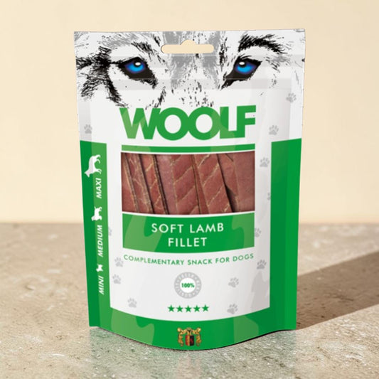 A stand-up pouch of WOOLF Soft Lamb Fillet dog treats, featuring a white and green package design with a wolf graphic and a clear window showing the lamb fillets inside, set on a stone surface against a beige background.