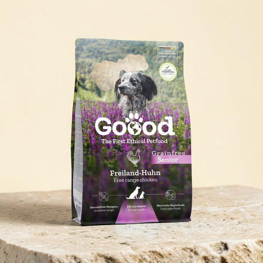 A bag of Goood grainfree senior Freiland-Huhn dog food, featuring a black and white dog in a purple flower field, standing on a stone surface against a beige background.