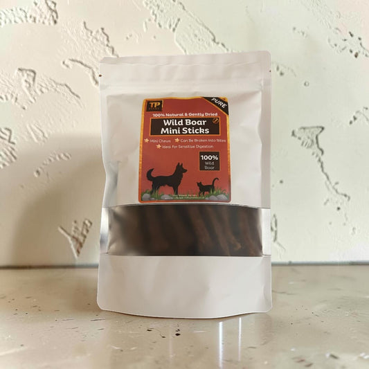 A stand-up pouch of TP Natural & Organic Wild Boar Mini Sticks dog treats, featuring a clear window showing the dark brown sticks inside, set against a textured off-white wall.