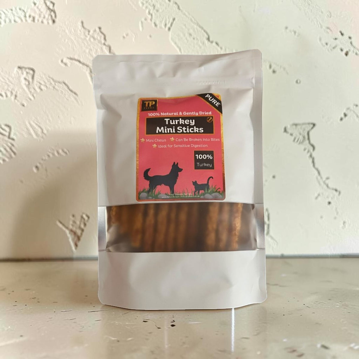 A stand-up pouch of TP Pure Turkey Mini Sticks dog treats, featuring a clear window that shows the brown sticks inside, set against a textured off-white wall.