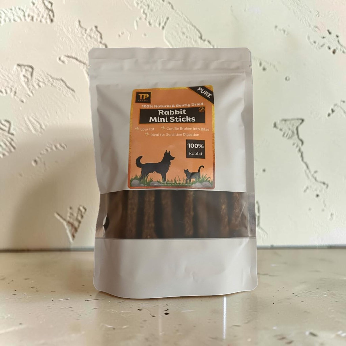 A stand-up pouch of TP Pure Rabbit Mini Sticks dog treats, featuring a white bag with a clear window showing the brown sticks inside, set against a textured off-white wall.