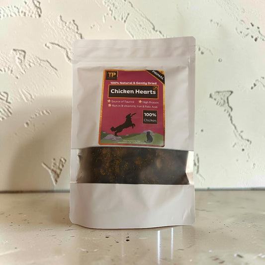 A stand-up pouch of TP Chicken Hearts dog treats, featuring a white bag with a clear window showing the dark brown treats inside, set against a textured off-white wall.
