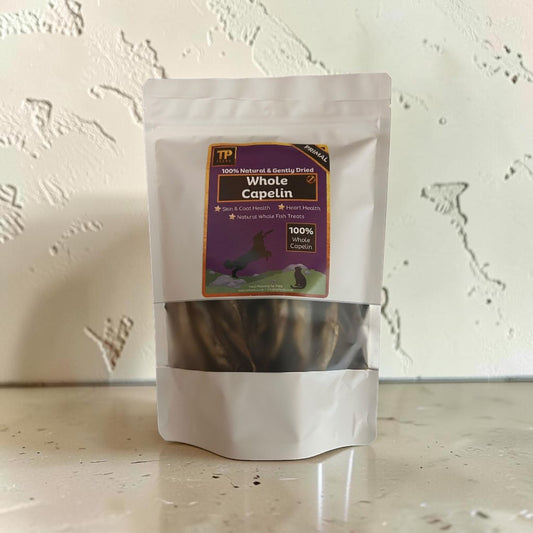 A stand-up pouch of TP Whole Capelin dog treats, featuring a white resealable bag with a clear window showing the dark brown treats inside, set against a textured off-white wall.