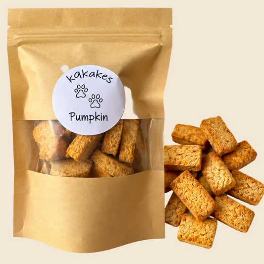 A stand-up kraft paper pouch with a clear window showing rectangular pumpkin-flavored treats inside, with several treats spilled beside it on a light cream background. A round white label on the pouch reads 'K9kakes Pumpkin' with paw prints.