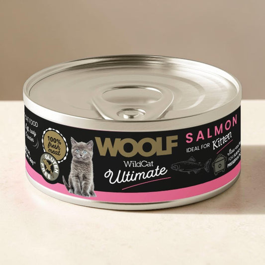 A can of Wolf WildCat Ultimate Salmon wet cat food, featuring a black and pink label with a grey cat illustration, set on a light beige surface against a neutral beige background.