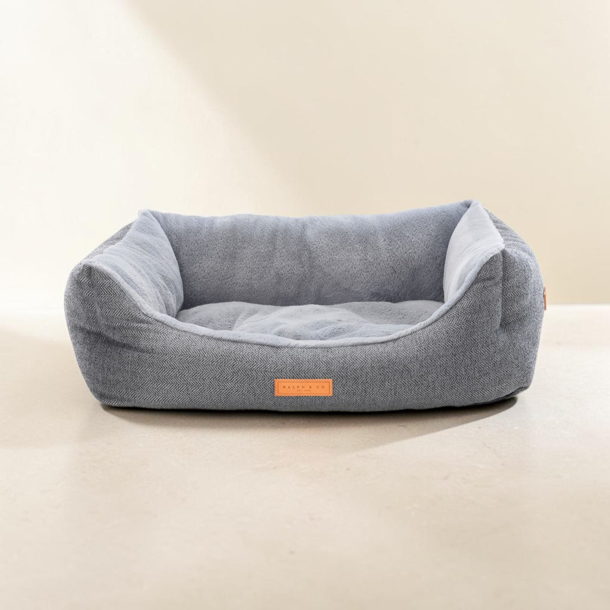 A light gray textured fabric pet bed with a plush gray interior cushion, shown on a neutral beige background. A small orange label on the front reads 'PUPPY HUT'.