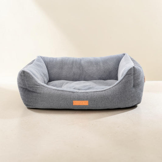 A light gray textured fabric pet bed with a plush gray interior cushion, shown on a neutral beige background. A small orange label on the front reads 'PUPPY HUT'.