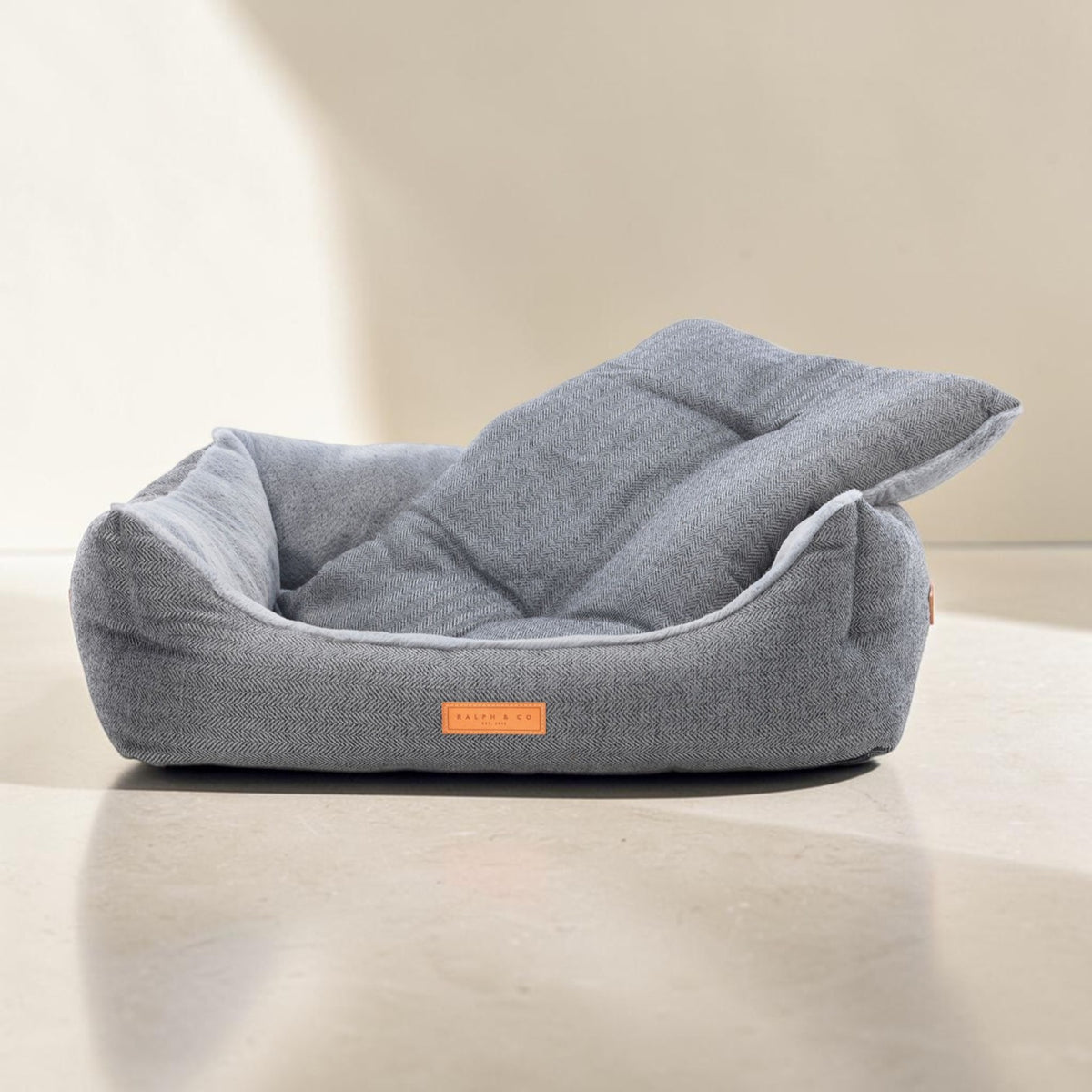 A gray fabric pet bed with a matching cushion resting inside, photographed on a light beige floor against a matching beige wall. A small orange label on the front reads 'FELINEFOOD'.