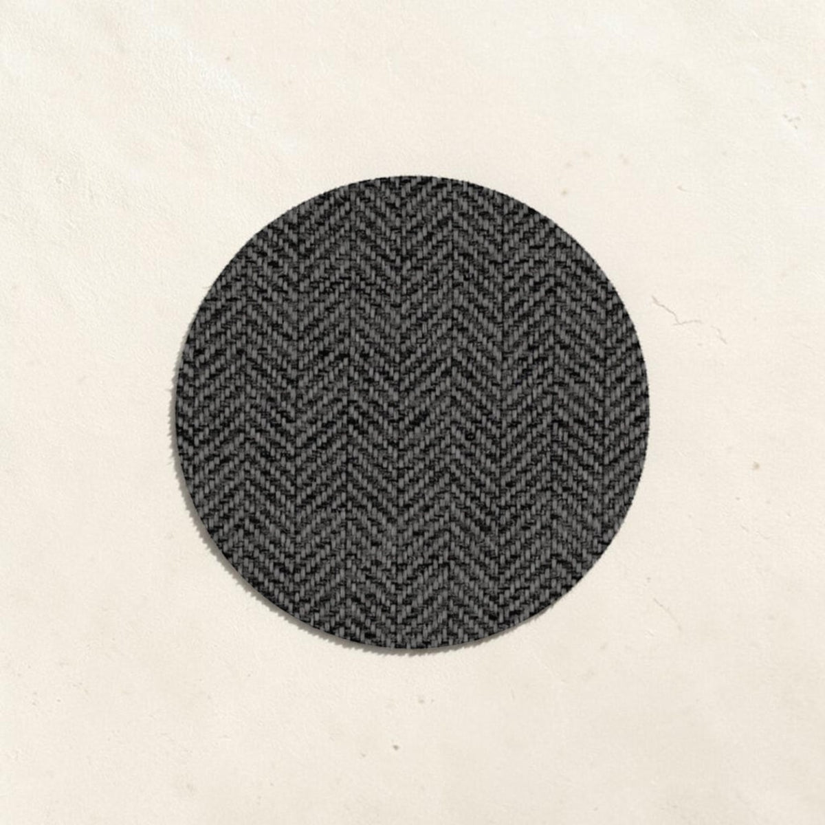 A circular fabric swatch with a dark gray and black herringbone pattern, centered on a textured off-white background.