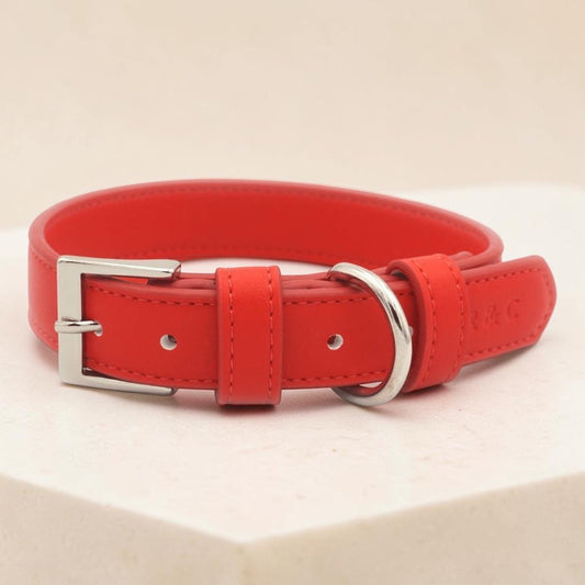 A bright red leather dog collar with a silver-tone metal buckle and D-ring, displayed on a light beige surface.