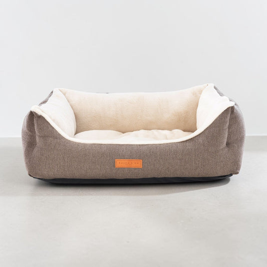 A rectangular pet bed with a textured taupe-brown fabric exterior, a cream fleece interior, and a small orange brand label on the front, shown on a light gray floor against a white wall.