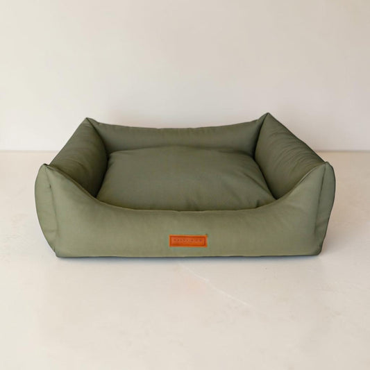 An olive green rectangular dog bed with a raised padded border and a small orange label on the front, photographed against a plain off-white wall and floor.