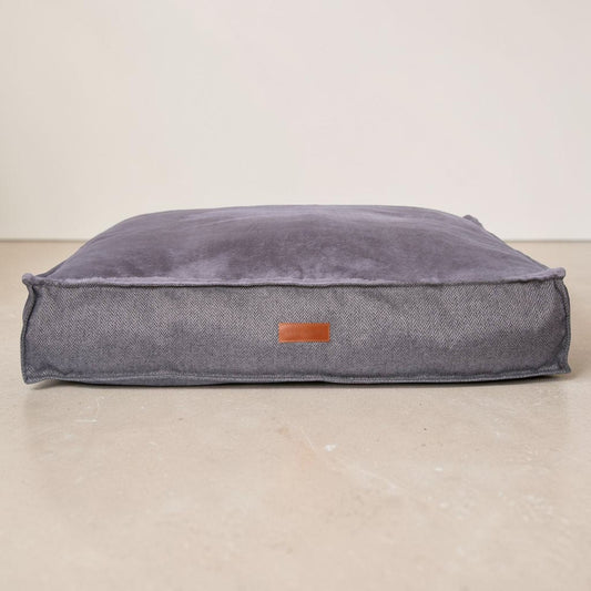 A square pet bed in a textured gray fabric with a plush top cushion, featuring a small brown leather brand patch on the front, set against a neutral beige background.