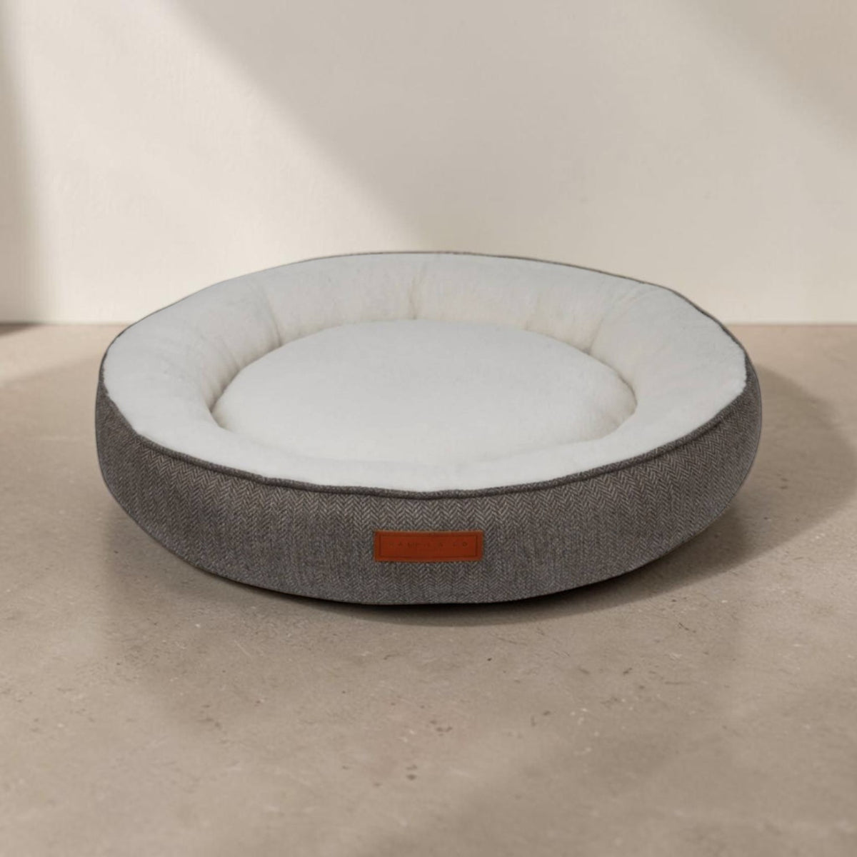 A round pet bed with a textured gray fabric exterior and a soft white interior, sitting on a neutral concrete surface against a beige wall.