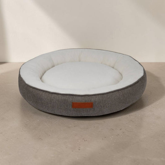 A round pet bed with a textured gray fabric exterior and a soft white interior, sitting on a neutral concrete surface against a beige wall.