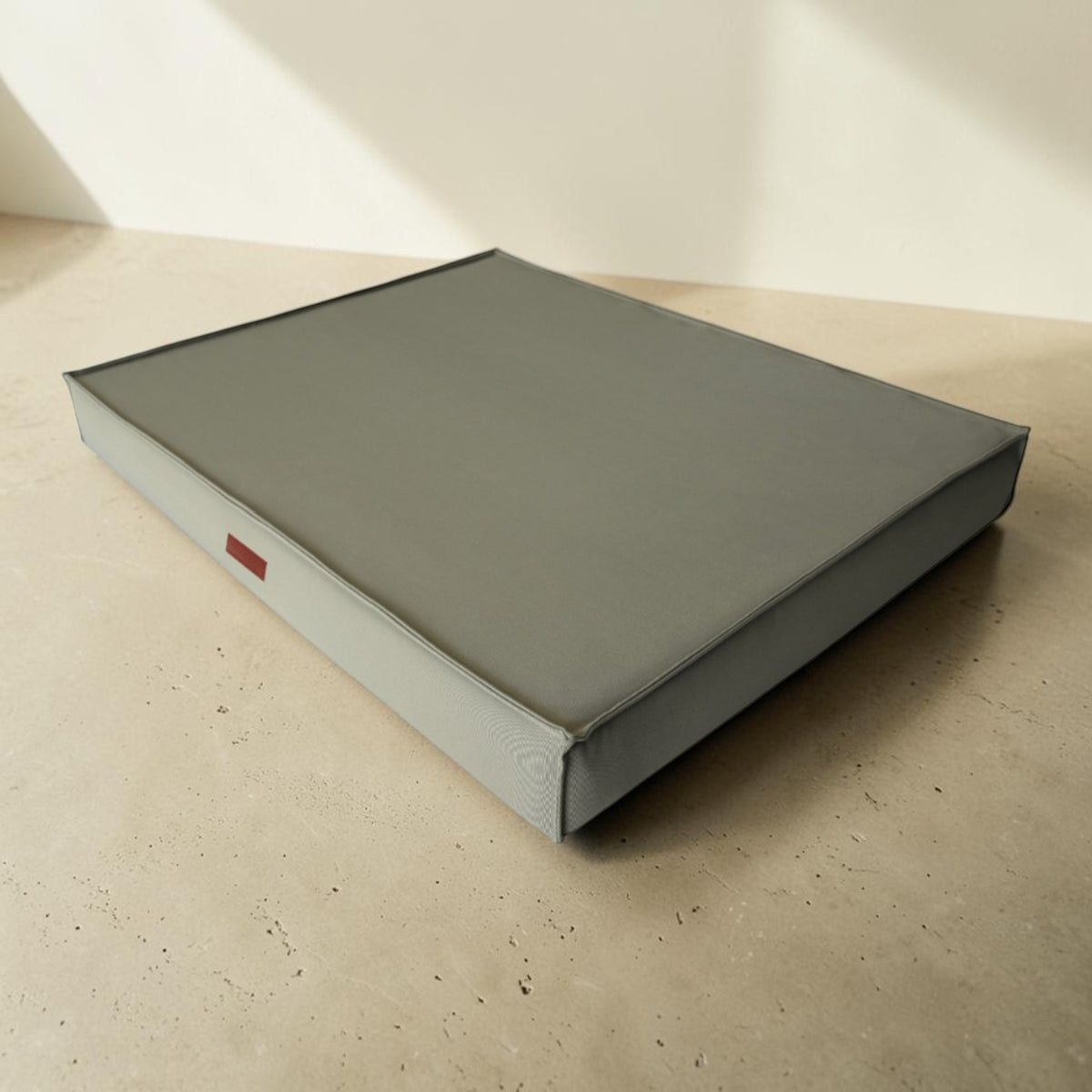A square gray upholstered bench or ottoman with a light gray side base and a small red rectangular accent on the side, photographed in a minimalist beige room setting.