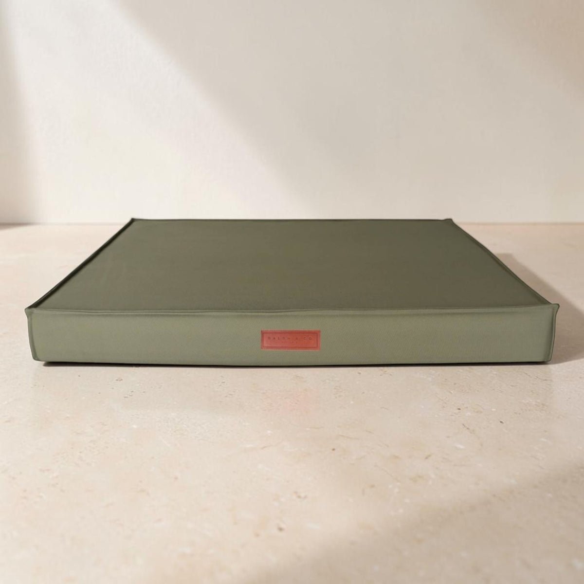 A square olive green hardcover book or album with a small red label on the front, resting on a light beige stone surface against a white wall.