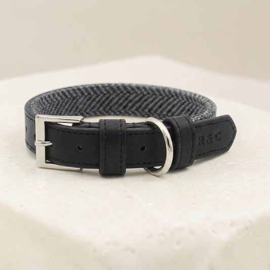 A black leather dog collar with a silver-tone buckle and metal ring, featuring a black and gray herringbone pattern on the underside, displayed on a textured cream surface.