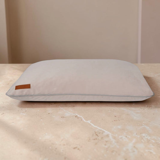A light beige rectangular pillow with a small brown leather brand tab on the left side, resting on a beige marble surface against a neutral beige background.