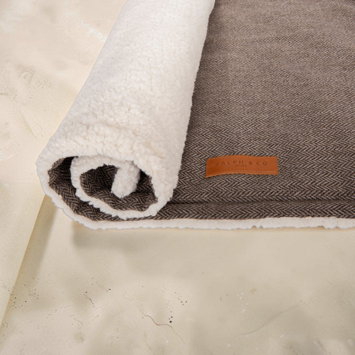 A rolled-up area rug showing its dual texture: a warm brown herringbone wool top and a plush off-white sherpa fleece underside, with a small tan leather brand tag reading 'The Rug Co.' on the side, set against a light beige stone surface.