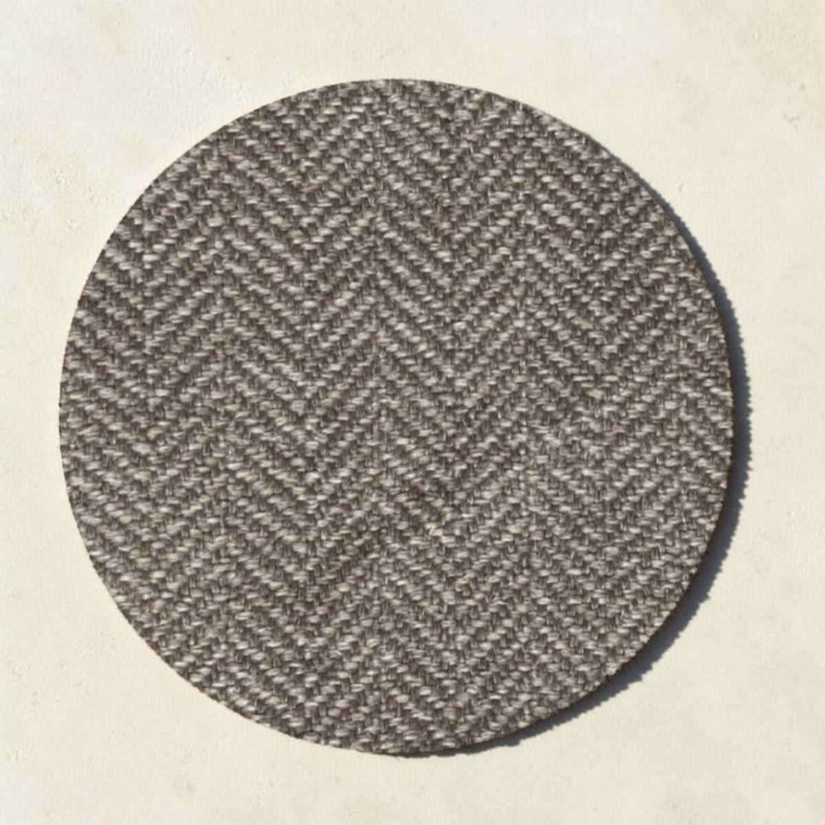 A round area rug with a brown and beige herringbone pattern, shown from a top-down view on a light beige textured background.