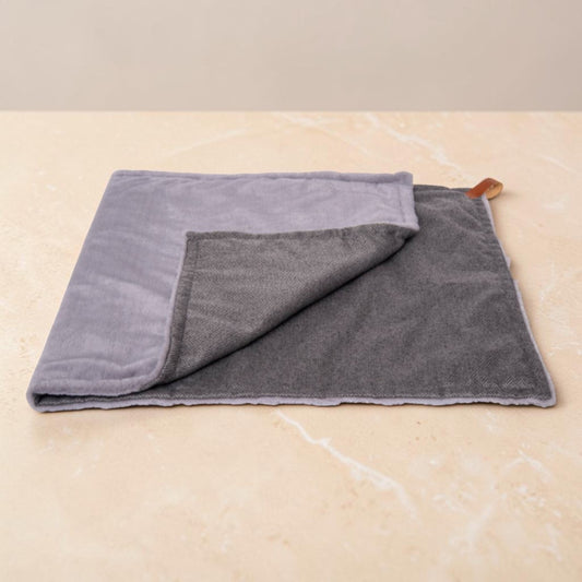 A folded gray microfiber cloth with a lighter gray reverse side and a small brown leather loop, displayed on a beige marble surface.