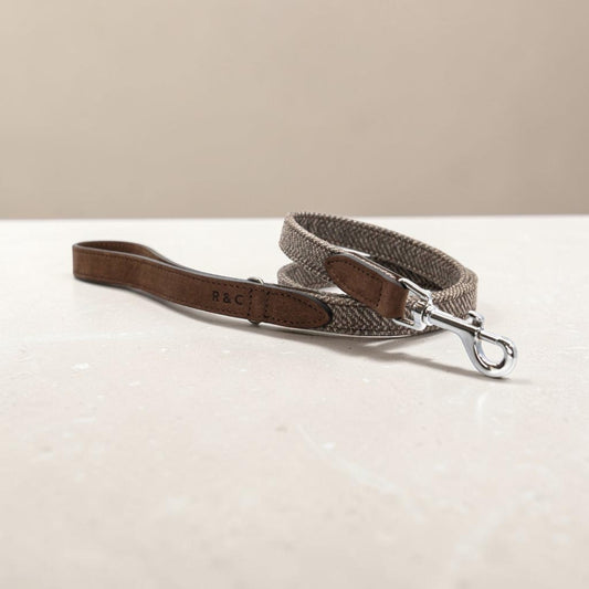 A dog leash with a brown leather handle and a woven gray and white herringbone strap, featuring a silver metal snap hook and 'R&C' branding on the leather.