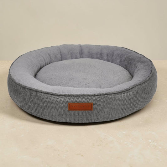 A round gray herringbone pet bed with a plush gray interior and a small brown leather brand patch, set against a neutral beige background.