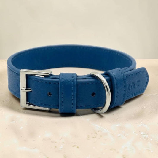 A royal blue leather dog collar with a silver-tone metal buckle and D-ring, displayed on a light beige textured surface.