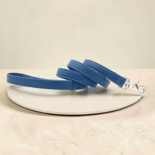 A blue faux-leather dog leash with silver metal hardware, coiled on a round white ceramic plate over a beige marble surface.