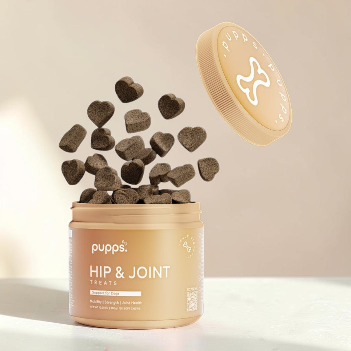 A beige plastic jar of Pups Hip & Joint Treats with the lid off, showing several dark brown heart-shaped treats floating out above the container on a light beige background.