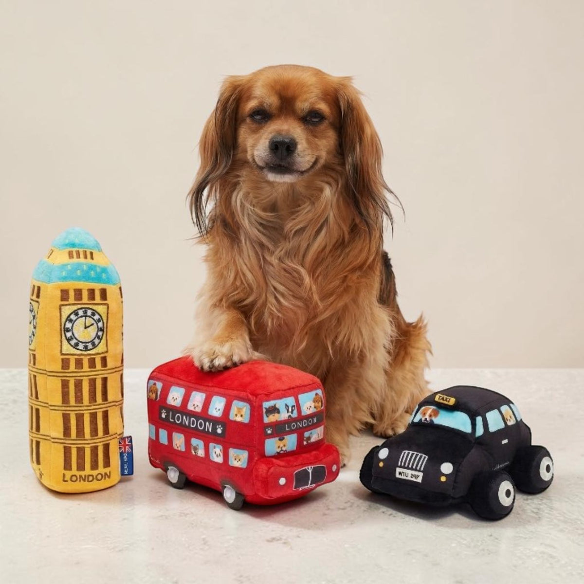 A small brown dog sits behind three London-themed plush toys on a light surface against a plain beige wall: a yellow tower toy labeled London, a red double-decker bus, and a black cab.